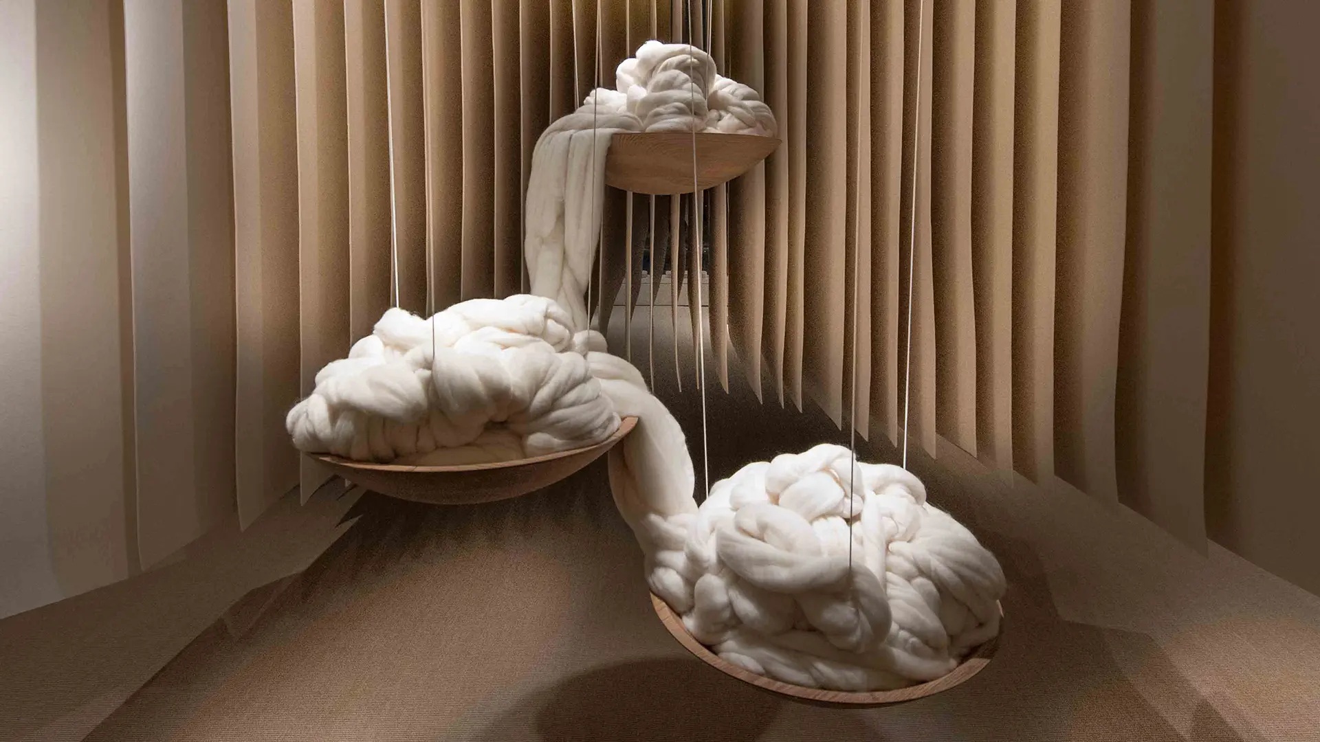 Large bundles of raw wool displayed in shallow wooden bowls, suspended at different heights within a sculptural installation space.